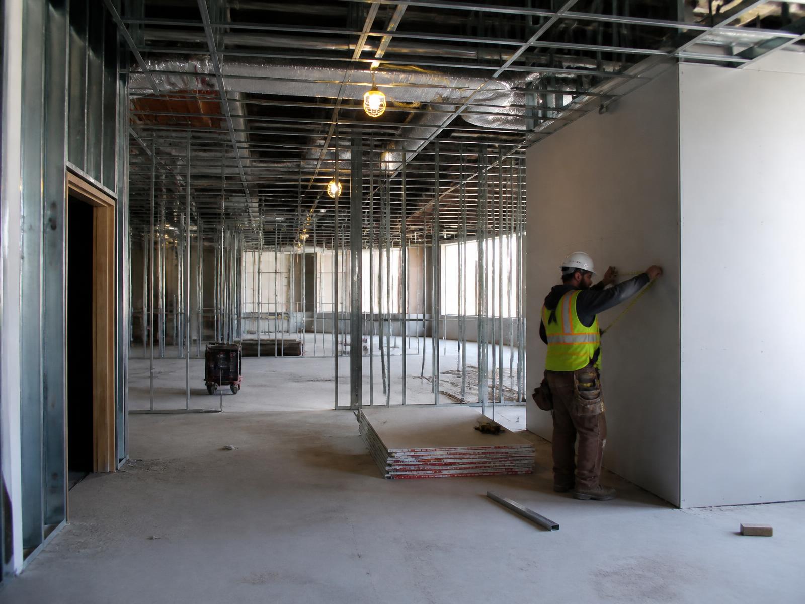Commercial interior build-out in progress with metal stud framing, exposed ductwork and a worker measuring drywall